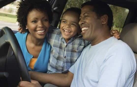 A smiling family sits together in a car, with the child snugly in the middle of the two adults in the front seat, enjoying peace of mind thanks to their uninsured motorist coverage.