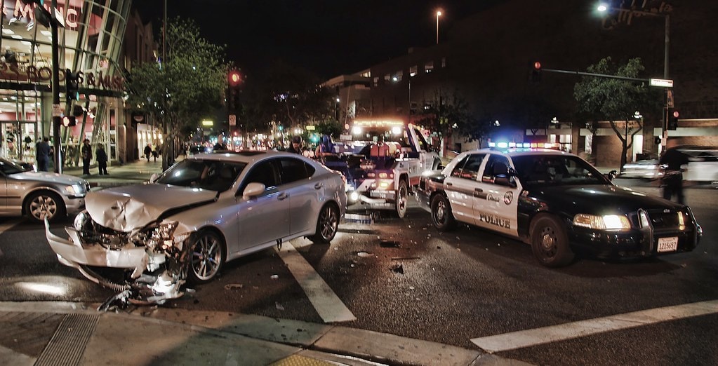 In a late-night incident at a city intersection, two cars, including a police vehicle, are damaged in what appears to be a drunk driving accident. The scene is illuminated by the flashing lights of a tow truck and police vehicles in the background.