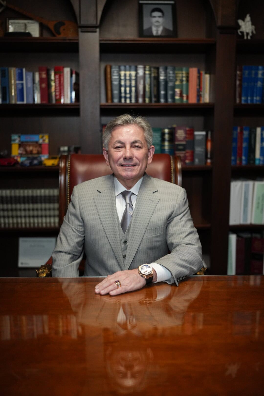 Person in a gray suit sitting at a wooden desk in front of a bookshelf filled with books and photos.