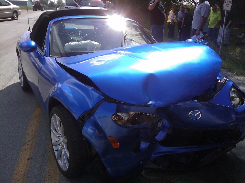 A blue convertible car with significant front-end damage is parked on the side of the road, suggesting a recent collision. People are gathered in the background, likely discussing the aftermath of this unfortunate event.