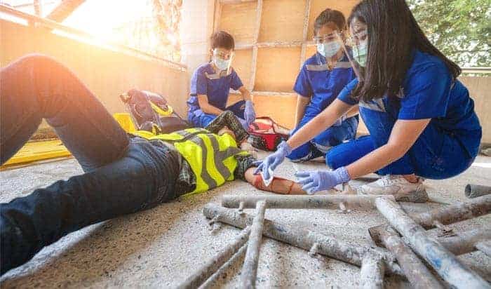 First responders swiftly aid a person in a high-visibility vest, likely involved in a construction accident, lying on the ground beside metal pipes. In Michigan, three masked and gloved rescuers efficiently attend to the situation with focused urgency.