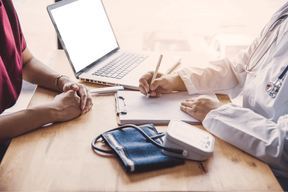 A doctor in a white coat writes on a clipboard while speaking with a patient, preparing for an independent medical exam. A blood pressure cuff and a laptop are on the wooden table.