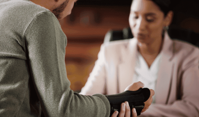 Two people sitting at a table, one wearing a gray sweater and holding a black wrist brace—a possible sign of Workers' Compensation needs—the other in a pink blazer.