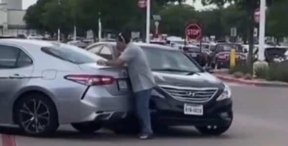 In a bustling parking lot, a person stands between a silver car and a black car, pushing them apart with caution, as if preventing an auto accident.