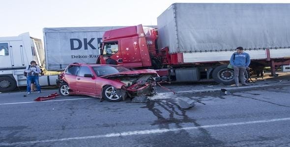 A red car with a severely damaged front sits in front of a large truck on the road, capturing the concern of two onlookers. As they assess the situation, thoughts might turn to Michigan Highway Accident Attorneys for expert guidance.