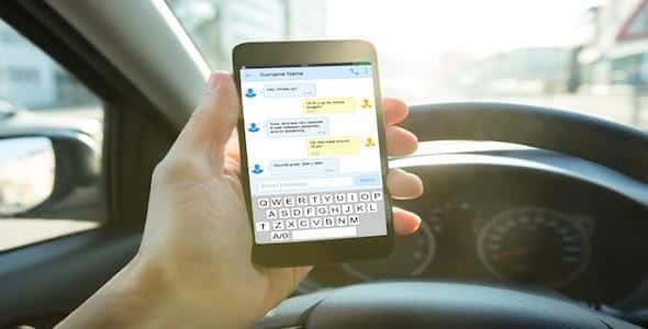 A person seated in a car is using a smartphone to display a chat conversation, with the steering wheel clearly visible, highlighting the potential for distracted driving accidents.