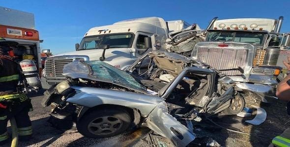 A mangled silver car is crushed between two large semi-trucks on a highway. Emergency responders are visible at the scene, highlighting the severity of the personal injury involved in this car rear-end truck accident.