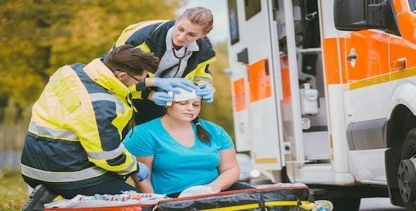 Paramedics assist a seated woman near an ambulance, tending to her with medical equipment after a car accident.