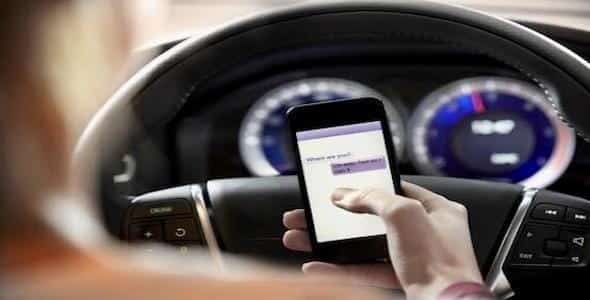 A person is holding a smartphone while driving, with the blurred car dashboard in the background, illustrating distracted driving.