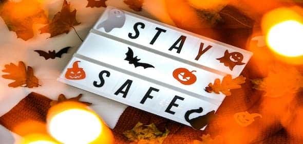 Illuminated sign with "Stay Safe" surrounded by autumn leaves and Halloween-themed decorations, including bats, pumpkins, and ghosts, offers a gentle reminder of Halloween safety tips for motorists and pedestrians, all under warm glowing lights.