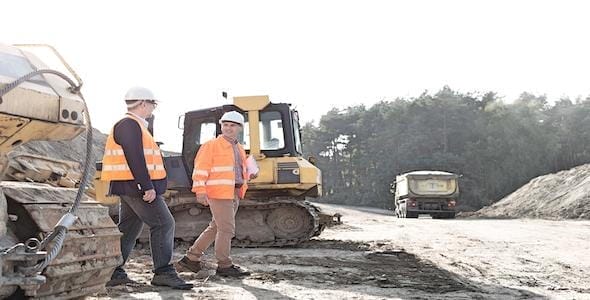 Two construction workers in reflective vests and helmets walk past machinery on a dirt road at a construction site, with a forest in the background. Safety is paramount here as they navigate their duties, mindful of potential risks highlighted by Michigan Heavy Equipment Accident Lawyers.