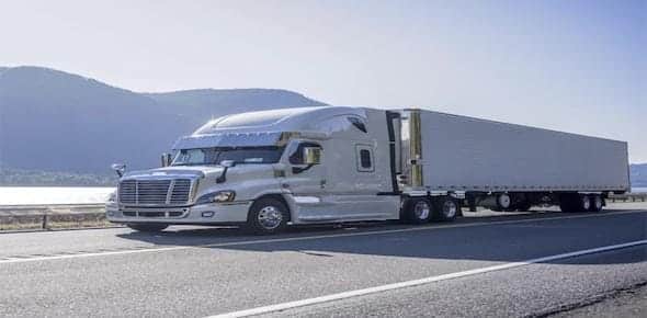 A silver semi-truck with a trailer is parked on a highway, framed by mountains and a shimmering body of water, highlighting the importance of understanding liability for semi-truck accidents in these stunning yet potentially hazardous locales.
