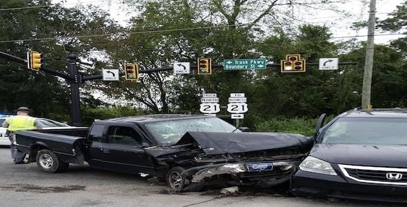 A black pickup truck and a black SUV are involved in a traffic accident at an intersection with traffic lights visible above. A person stands nearby on the left.