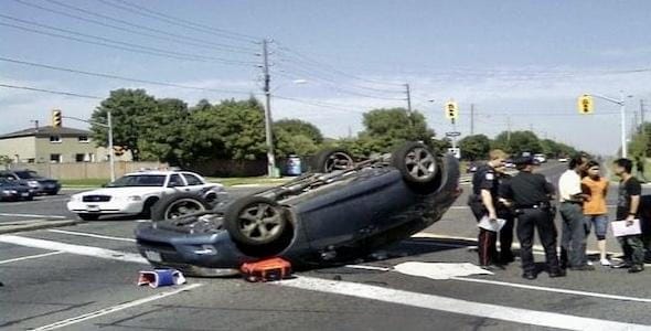 A rollover accident unfolded at a street intersection, leaving a car flipped on its roof. Police officers and concerned bystanders gathered as a patrol car and traffic lights loomed in the background.