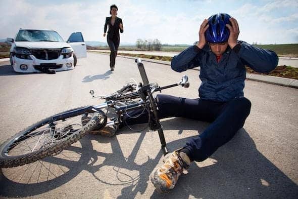 A cyclist sits on the road clutching their helmet by a fallen bicycle, indicating a bike accident, with a car and a person rushing towards them in the background.