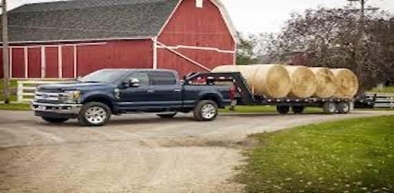 A black pickup truck hauls large round hay bales on a trailer past a red barn on a rural road.
