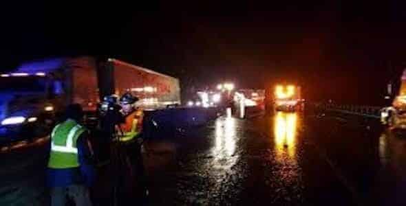 A nighttime highway scene unfolds with several trucks and emergency vehicles. Workers in reflective vests manage the situation, ensuring nighttime visibility on the wet road, hinting at recent rain. Truck drivers navigate cautiously as injury accident attorneys remain busy behind the scenes.