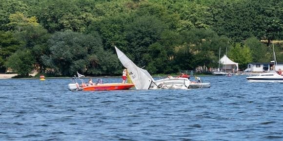 A small sailboat, capsized, is partially submerged in a lake as a motorboat with people nearby observes. Trees and a building are visible in the background, highlighting the importance of boating safety in such serene settings.