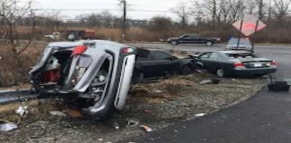 A Michigan car crash leaves three vehicles involved in an accident; one is overturned, while the other two are damaged. The scene is strewn with debris, a warning sign nearby, as authorities focus on determining fault.