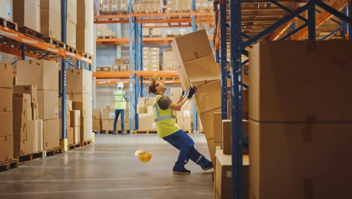 A worker in safety gear tries to stabilize a tumbling stack of boxes in a warehouse aisle, highlighting the importance of avoiding slip and fall accidents.