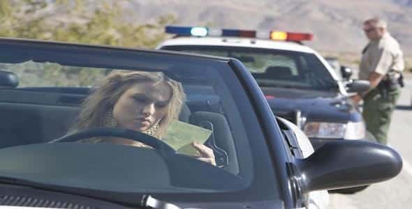 A woman in a convertible holds a document, possibly about what you need to know regarding insurance rates. In the background, a police officer stands next to a patrol car, suggesting she might be dealing with a distracted driving ticket.