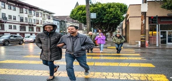 People walk across a crosswalk on a rainy day, with two individuals in front arm-in-arm while others follow behind. Buildings and trees line the street, offering a typical city scene where staying aware of pedestrian accident dos & don'ts is crucial for safety.