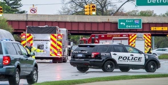 Police and heavy rescue vehicles, including government vehicles, are on the scene of an accident near a Michigan highway overpass with traffic signs and signals visible.