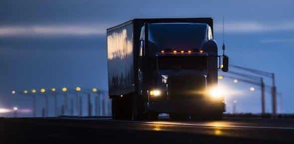 A large truck, piloted by a vigilant long haul truck driver with headlights on, travels down a highway at dusk, blurred streetlights painting the backdrop.