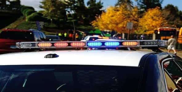 Police car with flashing lights at a roadside scene; other vehicles and people visible in the background on a sunny day with autumn trees, as authorities conduct a truck accident investigation.