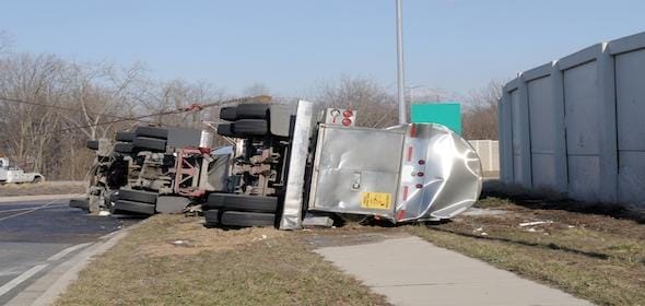 An overturned truck lies on the roadside, its wheels in the air due to shifting cargo. The toppled container rests near a concrete barrier and grassy area, a testament to the precarious journey of its truck driver.