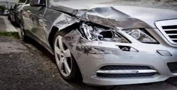 A silver car, seemingly the victim of a Michigan sideswipe accident, sits parked on the roadside with significant damage to its front left side, including a broken headlight and dented fender.