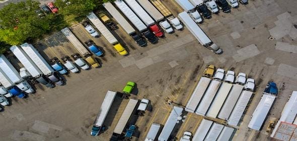 Aerial view of a trucking company lot with numerous parked semi-trucks and trailers in neat rows. Some trees and various colors of truck cabs are visible, showcasing the diverse fleet managed by experienced truck drivers.