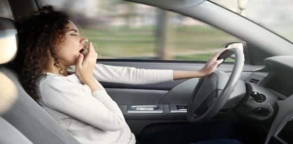 A person in the driver's seat yawns with one hand on the steering wheel, a clear sign of drowsy driving.