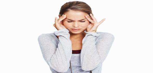 Woman with eyes closed and hands on temples, clearly suffering from a headache, possibly due to post-concussion syndrome. She's wearing a gray top against a white background, embodying the silent endurance of long-term pain.