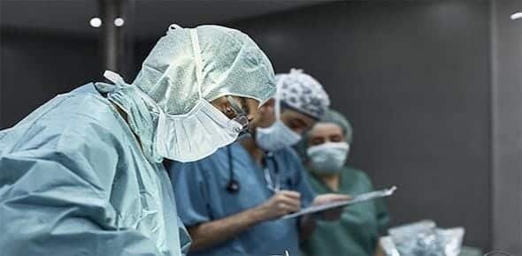 Doctors in scrubs and masks are meticulously performing surgery in an operating room, a procedure necessitated by a severe car accident. Two of them are diligently taking notes, ensuring every detail is accounted for during the operation.