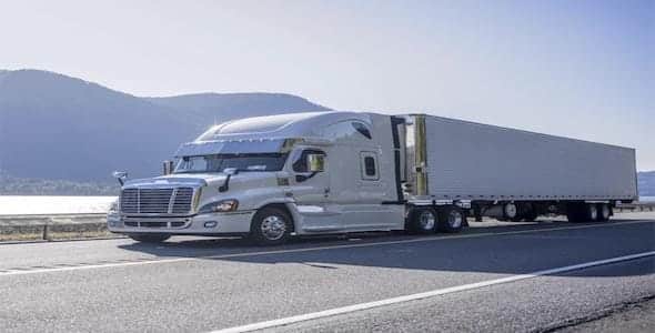 A white semi-truck with a long trailer navigates the highway, framed by mountains and a shimmering body of water in the background, evoking thoughts of trucking accident claims.