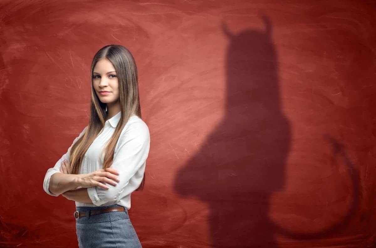 Woman with crossed arms in front of a red wall; her shadow, resembling an insurance adjuster, appears with horns and a tail.