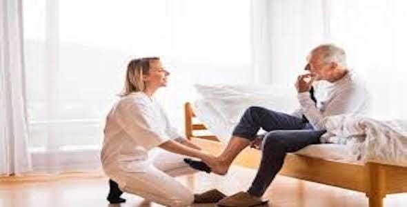 In a bright room, a caregiver gently assists an elderly man sitting on the bed, carefully placing a slipper on his foot. This tender moment reflects the essential support provided under Michigan's auto accident attendant care rate system.