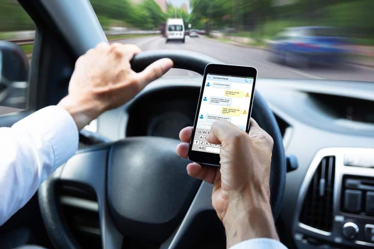 A person is texting on a smartphone while driving a new car. The steering wheel is in the left hand, and the road is visible through the windshield, raising concerns for the U.S. DOT as it sets fuel economy standards for safer driving practices.