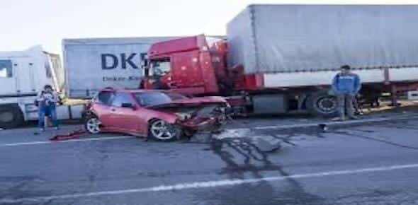 A red car with front-end damage is involved in a lane change accident with a large truck on the road. Two people stand nearby.
