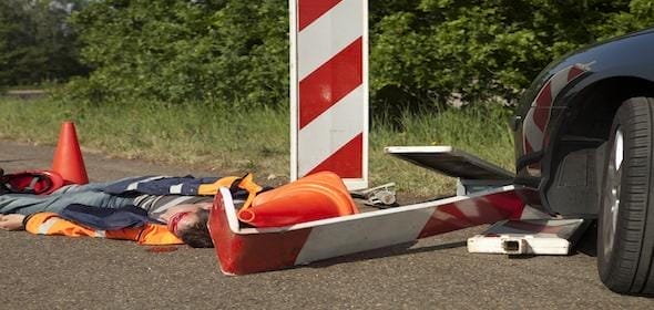 A person in a reflective vest lay next to a fallen road sign and traffic cones on a Michigan road beside a car, highlighting the risks involved in road construction zones.