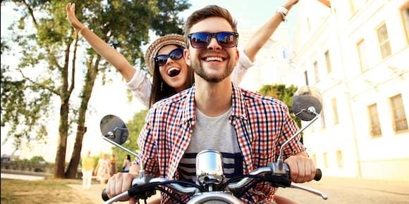 A man and woman ride a motorcycle, both smiling and wearing sunglasses. The woman raises her arms joyfully, embracing the thrill of the ride. Trees and buildings blur in the background, as they enjoy a carefree day without a hint of road rash or concern for motorcycle accidents.