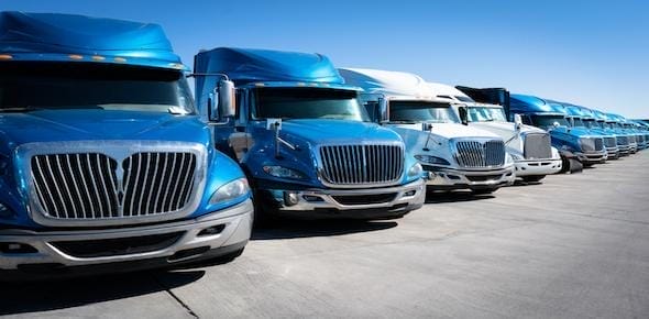A row of blue and white semi-trucks, owned by a reputable trucking company, sit parked under a clear blue sky.