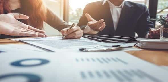Two people in business attire, possibly a motorcycle accident lawyer and a client, are discussing documents at a table filled with charts and a clipboard.