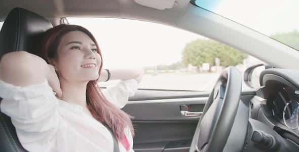 A person sits relaxed in an electric vehicle, hands behind their head, gazing out the window with a slight smile, as the autopilot smoothly navigates the road.