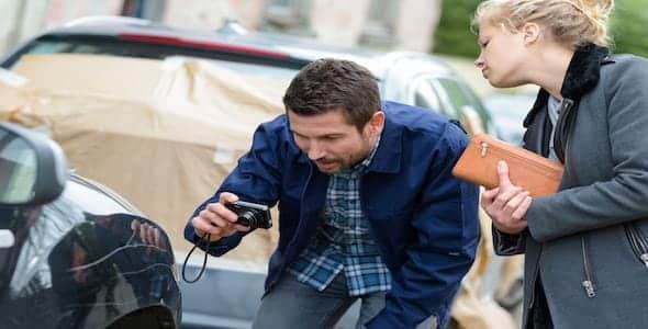 A man crouches to photograph the car at a crash scene, while a woman with a purse looks on. Cars with protective covers are in the background.