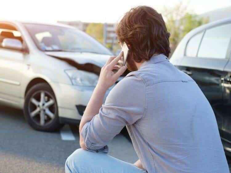 A man on the phone squats near two cars with front-end damage, possibly from a collision, in a parking area, contemplating car accident liability.