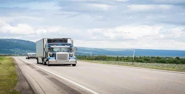 A blue semi-truck cruises along an open highway, framed by majestic mountains and cloudy skies. If you're dealing with truck driver negligence, consider a free consultation to navigate your next steps with ease.