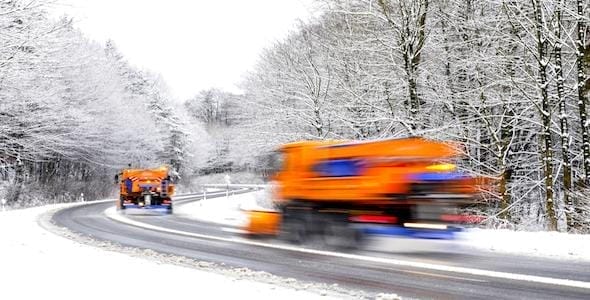 Two orange snowplows expertly clear a winding, snow-covered road bordered by snowy trees, ensuring safety and guarding against the need for snow plow accident attorneys.