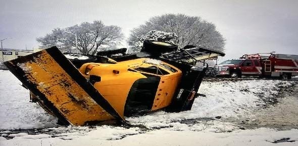 A utility truck accident left an overturned yellow snowplow in the snow, with a fire truck nearby and trees standing silent in the background.